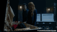 a man and a woman are standing next to a coffin with the website shadowhunterstv.com on the bottom
