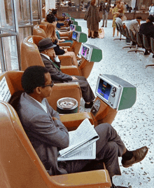 a man sits in a chair with a newspaper and a cup of coffee in front of a row of televisions