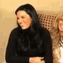 two women are sitting next to each other on a couch and smiling . one of the women is wearing a black sweater .