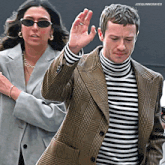 a man wearing a plaid jacket and a striped turtleneck stands next to a woman wearing sunglasses ..