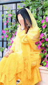 a woman in a yellow dress is covering her face with her hand while sitting in front of a bush of pink flowers .