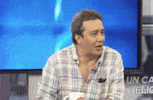 a man in a plaid shirt is sitting in front of a tv screen that says " un ca "