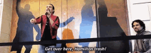 a man is singing into a microphone in front of a wall with a shadow of hamilton on it .
