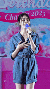 a woman in a denim jumpsuit sings into a microphone in front of a sign that says 2025