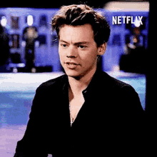 harry styles is wearing a black shirt and necklace while sitting in front of a netflix logo .
