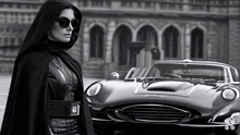 a black and white photo of a woman in a cape standing next to a car that says gt