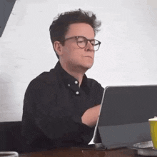 a man wearing glasses is sitting at a table using a laptop .