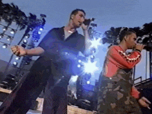 two men singing into microphones on a stage with a blue sky in the background