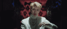 a man wearing a white suit and turtleneck is sitting in a red chair