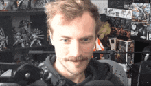 a man with a mustache looks at the camera in front of a sign that says ' a '
