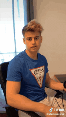a young man wearing a blue guess t-shirt is sitting in a chair