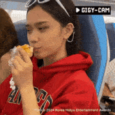 a woman in a red hoodie is eating a sandwich with a gigy-cam logo in the background