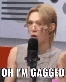 a man wearing glasses is talking into a microphone and says `` oh i 'm gagged '' .