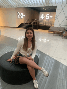 a woman sits on a black ottoman in front of a sign that says 2-3 4-5