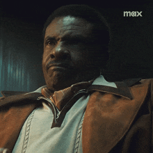 a man in a brown jacket and tie is sitting in a dark room with a max logo in the corner