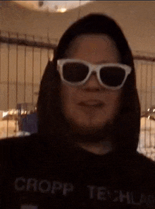 a woman wearing sunglasses and a black hoodie is standing in front of a fence .