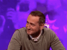 a man in a gray shirt is laughing while sitting in front of a purple background .