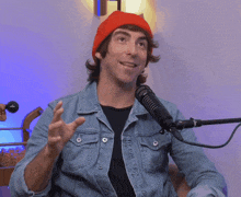 a man wearing a red beanie and denim jacket is talking into a microphone