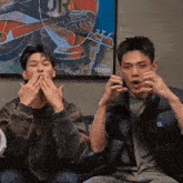 two young men are sitting next to each other on a couch covering their mouths .
