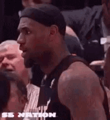 lebron james is sitting in the stands during a basketball game and talking to a referee .
