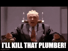 a man in a suit and tie is sitting in a chair and says i 'll kill that plumber