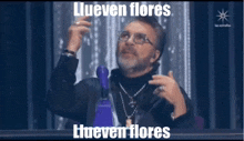 a man with glasses and a beard is sitting in front of a sign that says llueven flores on it