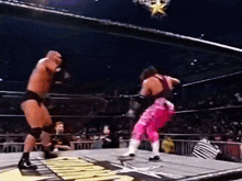 two men are wrestling in a ring with a referee watching . one of the men is wearing pink pants .