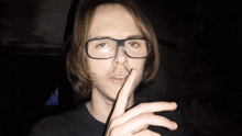 a man wearing glasses and a black shirt holds his finger to his lips