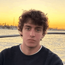 a young man with curly hair is sitting on a boat in front of the water .