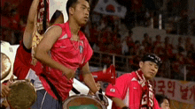 a man in a red shirt is holding a drum while standing in front of a crowd .