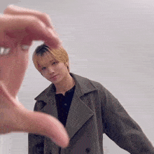 a person making a heart shape with their fingers in front of a young man in a trench coat