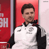 a man wearing a manchester united jacket sits in front of a red sign