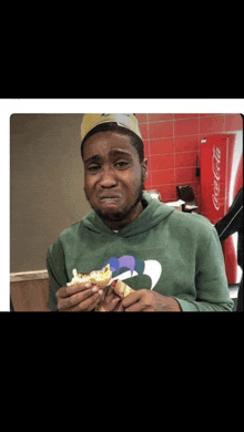 a man in a green hoodie is eating a sandwich in front of a coca-cola machine
