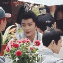 a man with a crown on his head stands in front of a bunch of red flowers