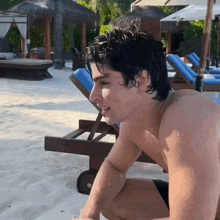 a shirtless man sits on a beach with a blue chair in the background