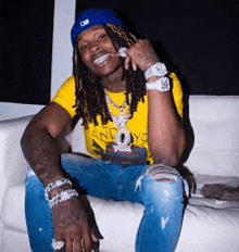 a man with dreadlocks is sitting on a white couch wearing a yellow shirt and a blue hat .