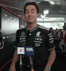 a man wearing a petronas shirt stands in front of a fox sports microphone