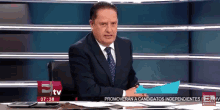 a man in a suit and tie is sitting at a desk in front of a screen that says etv