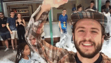 a man with a beard wearing a plaid shirt and a hat stands in front of a crowd of people