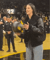 a woman in a black jacket is holding a basketball in front of a crowd with the number 22 behind her