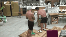 two men without shirts are cleaning a wooden floor with a mop in a kitchen with playplus.com in the corner