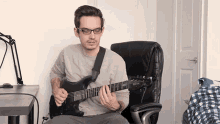 a man wearing glasses is sitting in a chair playing an electric guitar