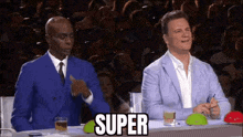 two men in suits are sitting at a table in front of a crowd and one of them is pointing at the word super .