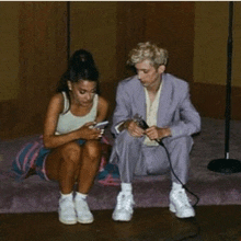 a man in a suit sits next to a woman who is sitting on a bed looking at her phone