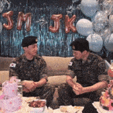two men in military uniforms are sitting on a couch in front of balloons that say jmjk
