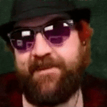 a man with a beard and sunglasses is wearing a hat .