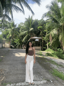 a woman in a brown tank top and white pants stands on the side of a road in front of palm trees