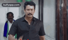 a man with a mustache and a black shirt is standing in a hallway and looking at the camera .