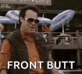 a man wearing sunglasses and a vest is standing in front of a building with umbrellas and says `` front butt '' .