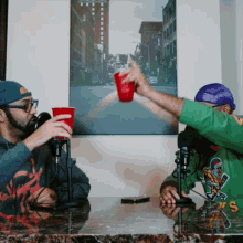 two men toasting with red cups in front of a painting of a city